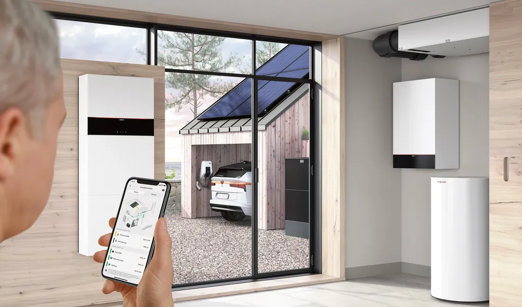 Viessmann ViCare App auf dem Smartphone, dahinter Inneneinheit der Wärmepumpe, Stromspeicher und ein Elektroauto, das unter einem Solarcarport lädt